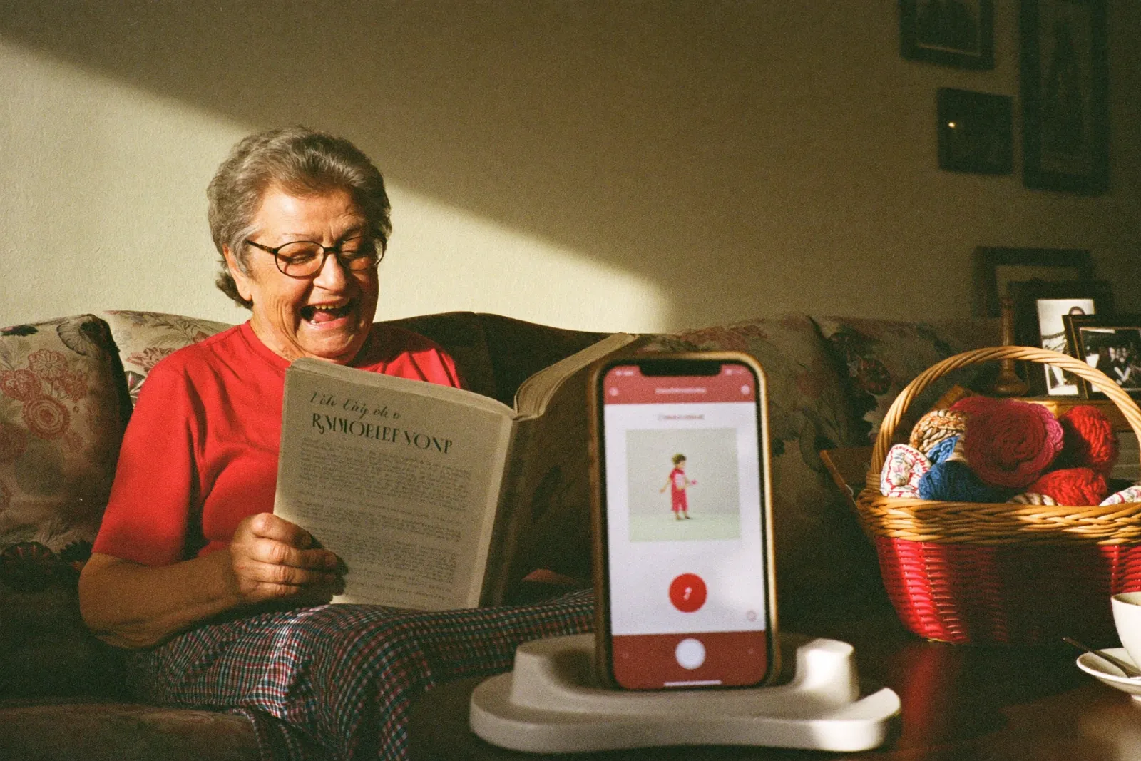 A grandmother on her sofa reading a picture book aloud to her phone, propped on the coffee table